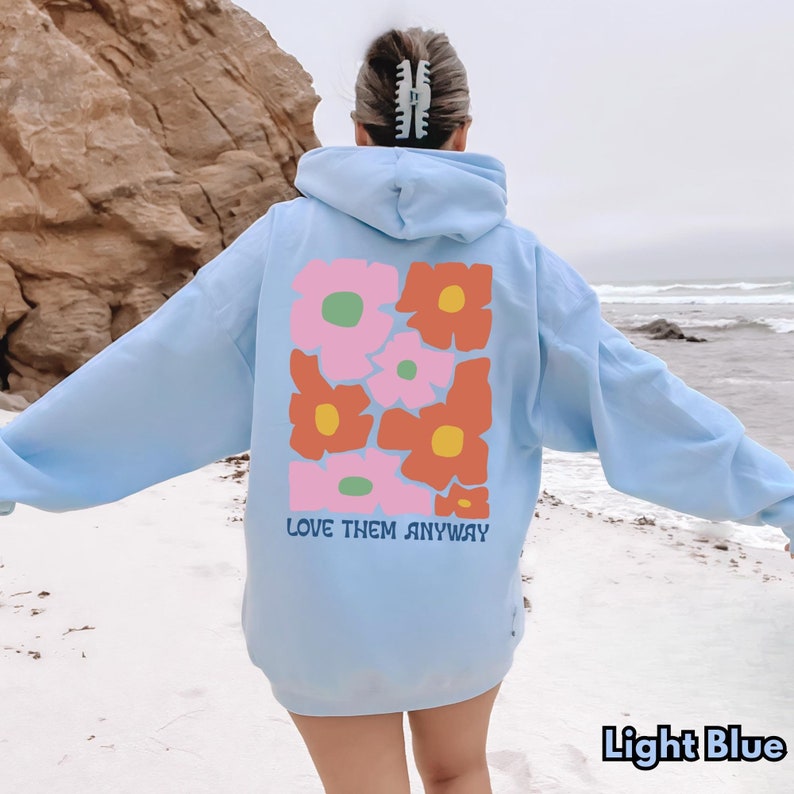 May include: A light blue hooded sweatshirt featuring a large graphic on the back with stylized pink and orange flowers with yellow and green centers. The text "LOVE THEM ANYWAY" is printed in dark blue letters below the floral design.
