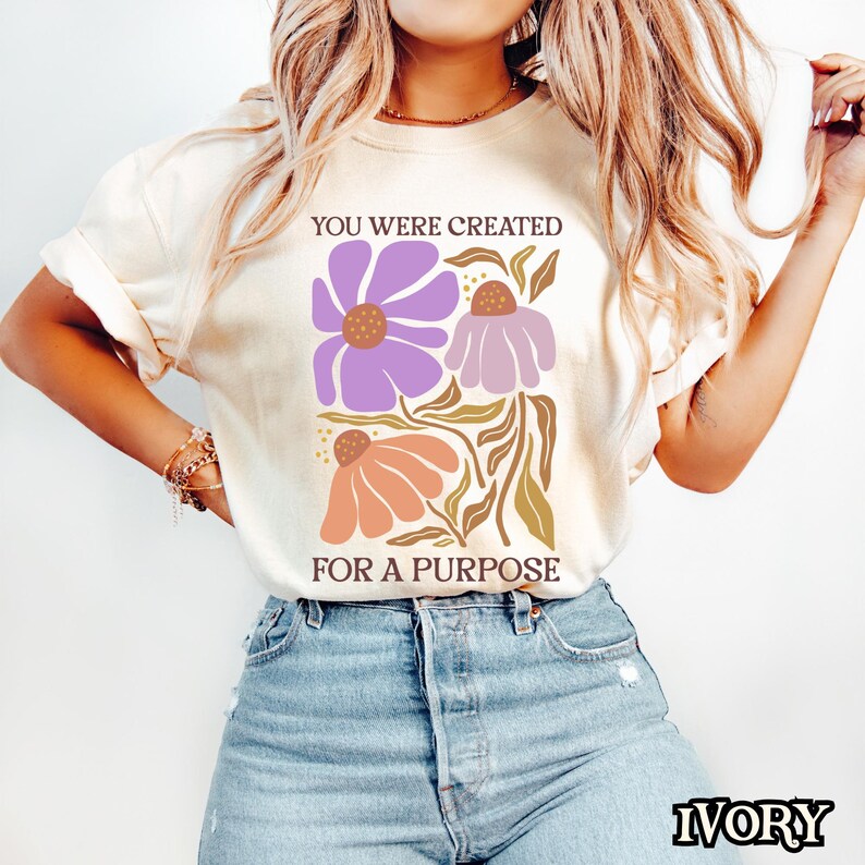 May include: A white t-shirt with the text "You were created for a purpose" printed on it. The text is in brown lettering and is surrounded by a floral design with purple, pink, and orange flowers.