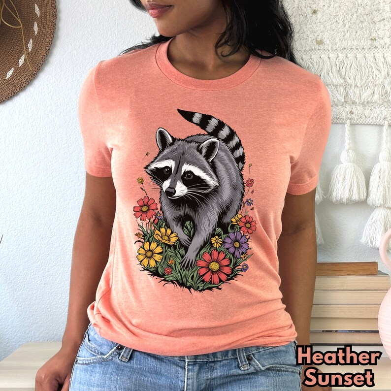 May include: A peach-colored t-shirt with a graphic of a raccoon walking through a field of colorful flowers.
