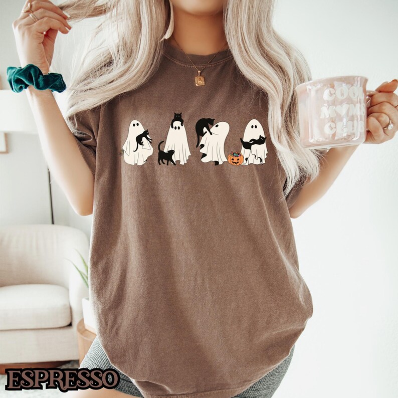 K&ouml;nnte beinhalten: Ein braunes T-Shirt mit einem Aufdruck von f&uuml;nf Geistern, die schwarze Katzen halten. Die Geister haben verschiedene Gesichtsausdr&uuml;cke, und einer h&auml;lt einen kleinen K&uuml;rbis. Ein Becher mit dem Text "Cool Mom Crew" ist im Hintergrund zu sehen.