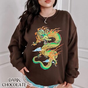 K&ouml;nnte beinhalten: Ein braunes Sweatshirt mit einem grafischen Aufdruck eines gr&uuml;nen und gelben Drachen mit orangefarbenen Flammen. Der Drache ist von wei&szlig;en Wolken umgeben.