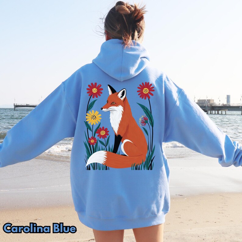 May include: Carolina Blue hoodie featuring a vibrant illustration of a fox amidst colorful flowers. The fox has orange fur, a white chest, and a fluffy white tail. The flowers are red and yellow. The hoodie is a soft, comfortable material.