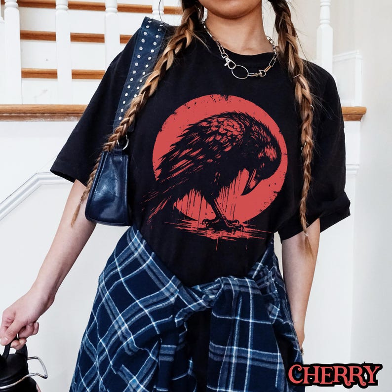 May include: Black t-shirt with a graphic of a black crow standing in a red circle. The crow is facing left and has its wings spread out. The t-shirt is being worn by a person with long, braided hair.