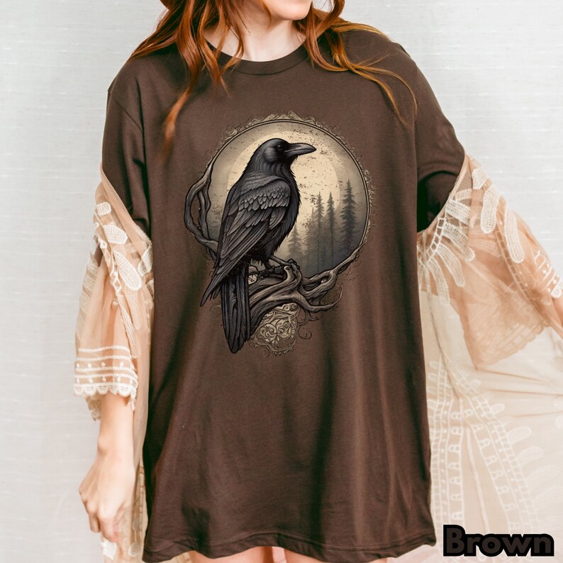 May include: A brown t-shirt with a graphic of a black raven perched on a branch in front of a moon and forest. The raven is detailed with a dark gray color and the background is a light brown.