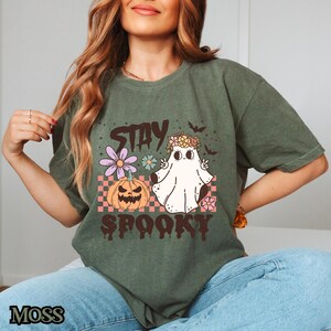 K&ouml;nnte beinhalten: Ein gr&uuml;nes T-Shirt mit einem Aufdruck eines Geistes mit einer Blumenkrone, einem K&uuml;rbis und dem Text "Stay Spooky".
