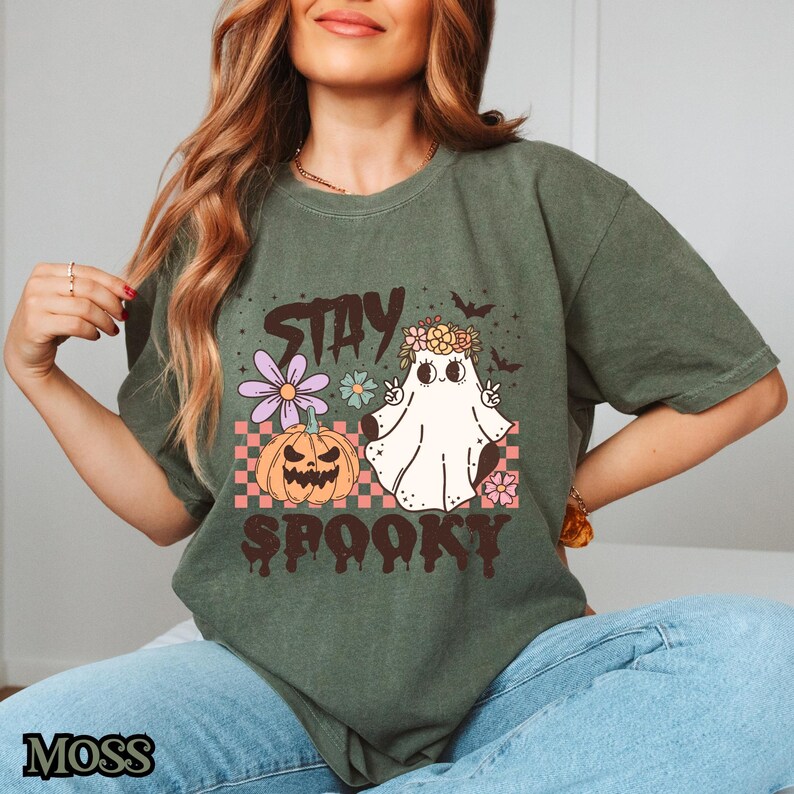 K&ouml;nnte beinhalten: Ein gr&uuml;nes T-Shirt mit einem Aufdruck eines Geistes mit einer Blumenkrone, einem K&uuml;rbis und dem Text "Stay Spooky".