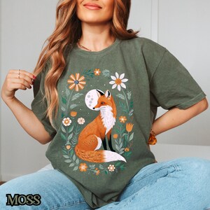 Op de afbeelding: Een groen T-shirt met een afbeelding van een vos die in een bloemenveld zit. De vos is oranje en wit, en de bloemen zijn in verschillende kleuren. De maan is zichtbaar op de achtergrond.