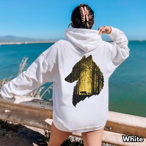 May include: A white hooded sweatshirt featuring a large graphic on the back. The graphic is a silhouette of a wolf head filled with a yellow-toned forest scene, depicting tall trees and two small animals walking through the woods.