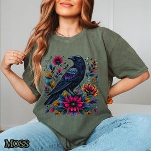 May include: A green t-shirt with a black raven and colorful floral design. The raven is perched on a branch with flowers in shades of pink, purple, and blue.