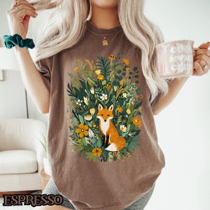 May include: A brown t-shirt with a graphic of a fox sitting in a field of flowers. The fox is orange and white, and the flowers are yellow, pink, and green.