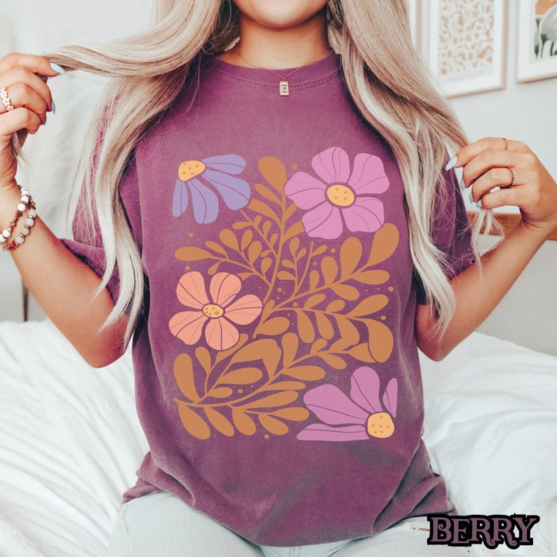 Op de afbeelding: Een paars T-shirt met een bloemenontwerp met roze, paarse en oranje bloemen met bruine stelen en bladeren.