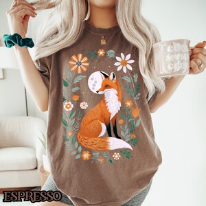 Op de afbeelding: Een bruin T-shirt met een afbeelding van een vos die in een cirkel van bloemen en bladeren zit. De vos is oranje en wit, en de bloemen zijn in tinten roze, geel en groen. De maan is zichtbaar op de achtergrond.