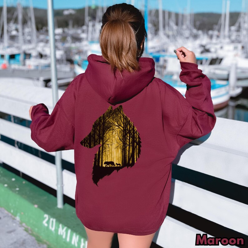 May include: A maroon hooded sweatshirt featuring a large graphic on the back. The graphic is a silhouette of a wolf head filled with a yellow-toned forest scene, showing two smaller wolves walking among the trees. The hoodie has long sleeves.