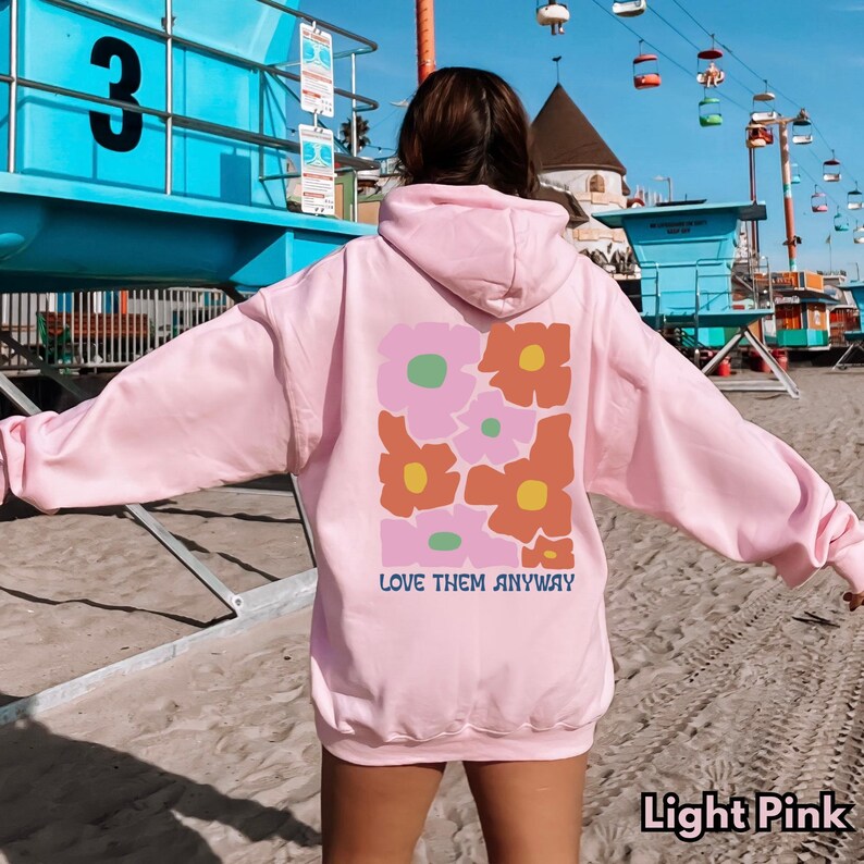 May include: A light pink hooded sweatshirt featuring a large graphic on the back with stylized flowers in shades of pink, orange, and green. Below the floral design, the text reads LOVE THEM ANYWAY in blue letters. The hoodie has long sleeves.