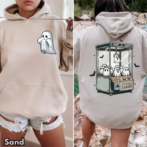 Pode incluir: Um moletom bege com um gr&aacute;fico de fantasma pixelado na frente e uma m&aacute;quina de garra com fantasmas dentro nas costas. O texto "Grab a Ghost" est&aacute; impresso na m&aacute;quina de garra. O moletom &eacute; modelado por uma pessoa que usa shorts jeans.