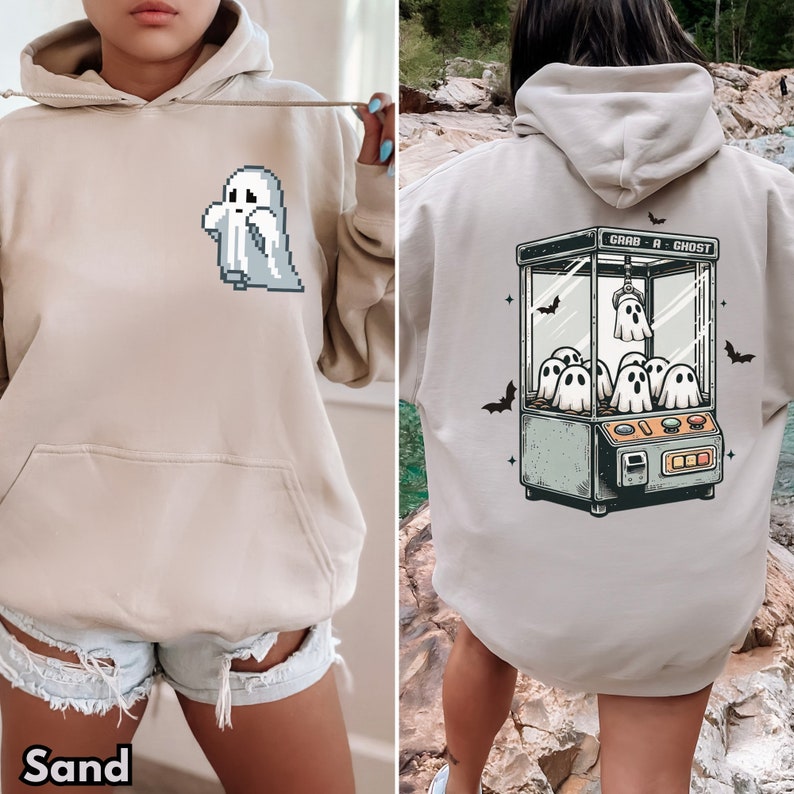 Pode incluir: Um moletom bege com um gr&aacute;fico de fantasma pixelado na frente e uma m&aacute;quina de garra com fantasmas dentro nas costas. O texto "Grab a Ghost" est&aacute; impresso na m&aacute;quina de garra. O moletom &eacute; modelado por uma pessoa que usa shorts jeans.
