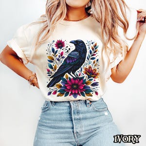 May include: A white t-shirt with a colorful floral design featuring a black raven. The raven is perched on a branch with pink, blue, and yellow flowers.