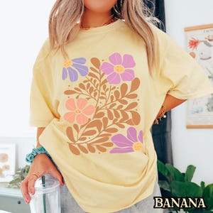 Op de afbeelding: Een geel T-shirt met een bloemenprint met paarse, roze en oranje bloemen met bruine stelen en bladeren.