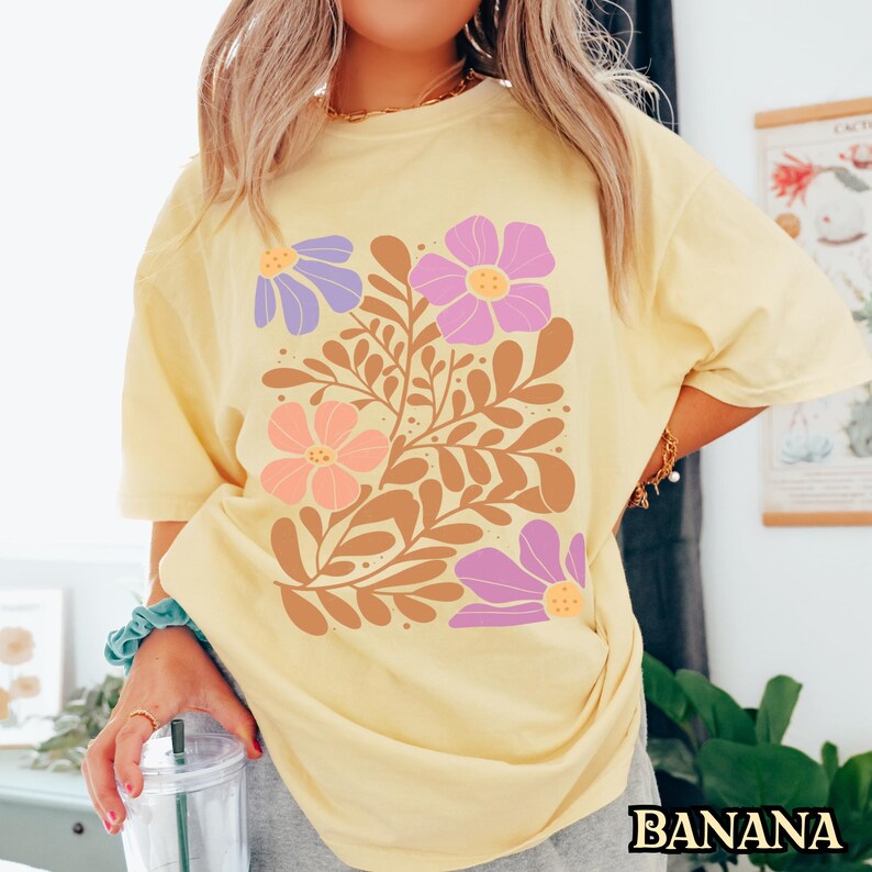 Op de afbeelding: Een geel T-shirt met een bloemenprint met paarse, roze en oranje bloemen met bruine stelen en bladeren.