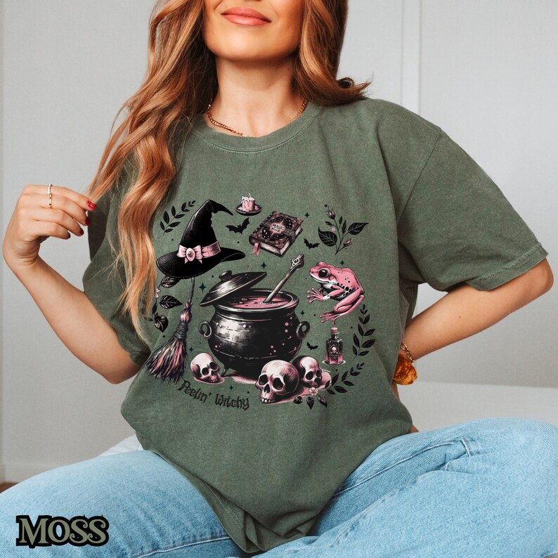 May include: Green t-shirt with a witchy graphic featuring a cauldron, a frog, a witch's hat, a book, a broom, and skulls. The text "Feelin' Witchy" is printed on the shirt.