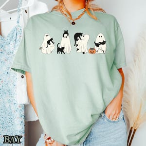 K&ouml;nnte beinhalten: Ein hellgr&uuml;nes T-Shirt mit einem Aufdruck von schwarzen Katzen und Geistern. Die Katzen tragen Hexenh&uuml;te und halten K&uuml;rbisse. Der Text "BOO" ist auf dem T-Shirt gedruckt.
