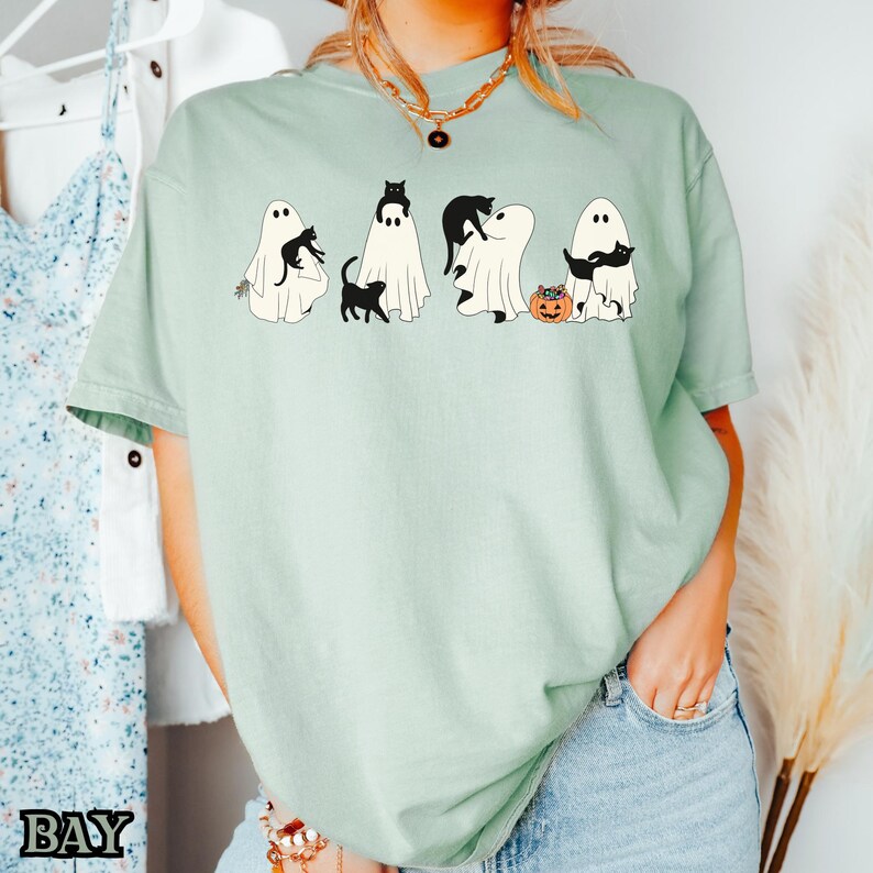 K&ouml;nnte beinhalten: Ein hellgr&uuml;nes T-Shirt mit einem Aufdruck von schwarzen Katzen und Geistern. Die Katzen tragen Hexenh&uuml;te und halten K&uuml;rbisse. Der Text "BOO" ist auf dem T-Shirt gedruckt.