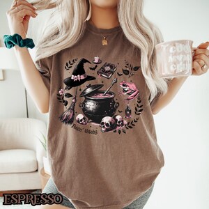 May include: Brown t-shirt with a pink and black witchy design featuring a cauldron, a witch's hat, a frog, a broom, and skulls. The text "Feelin' Witchy" is printed on the shirt.