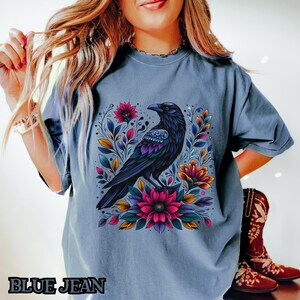 May include: A blue jean t-shirt with a graphic of a black crow with colorful floral accents. The crow is perched on a branch with flowers and leaves.