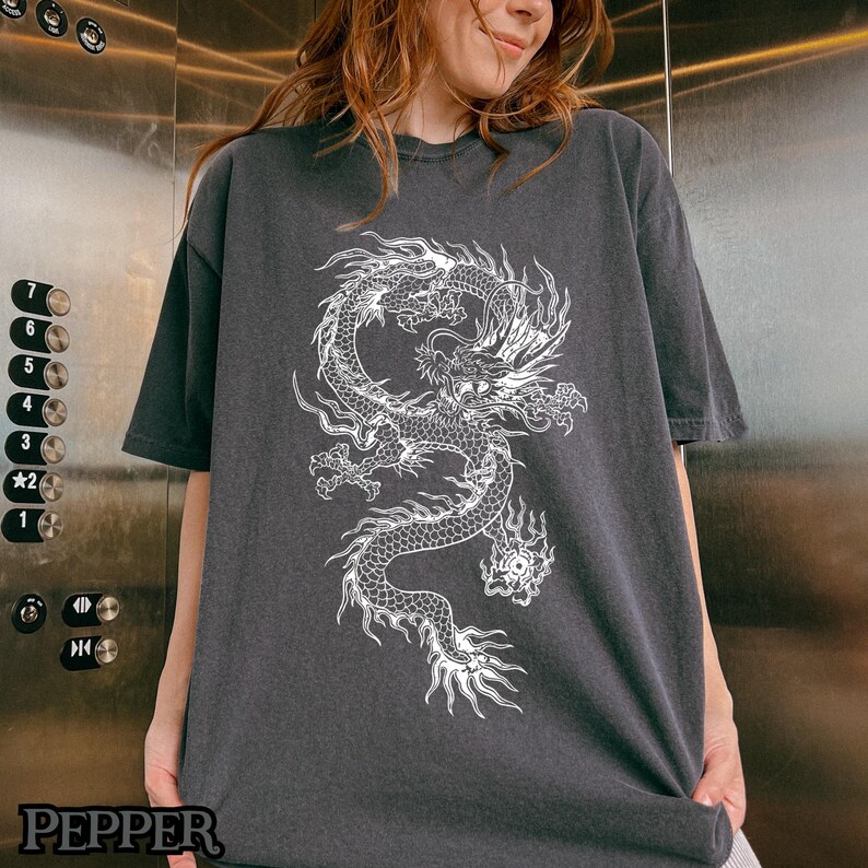 May include: A dark gray t-shirt with a white graphic of a dragon. The dragon is detailed with scales and has a fierce expression.