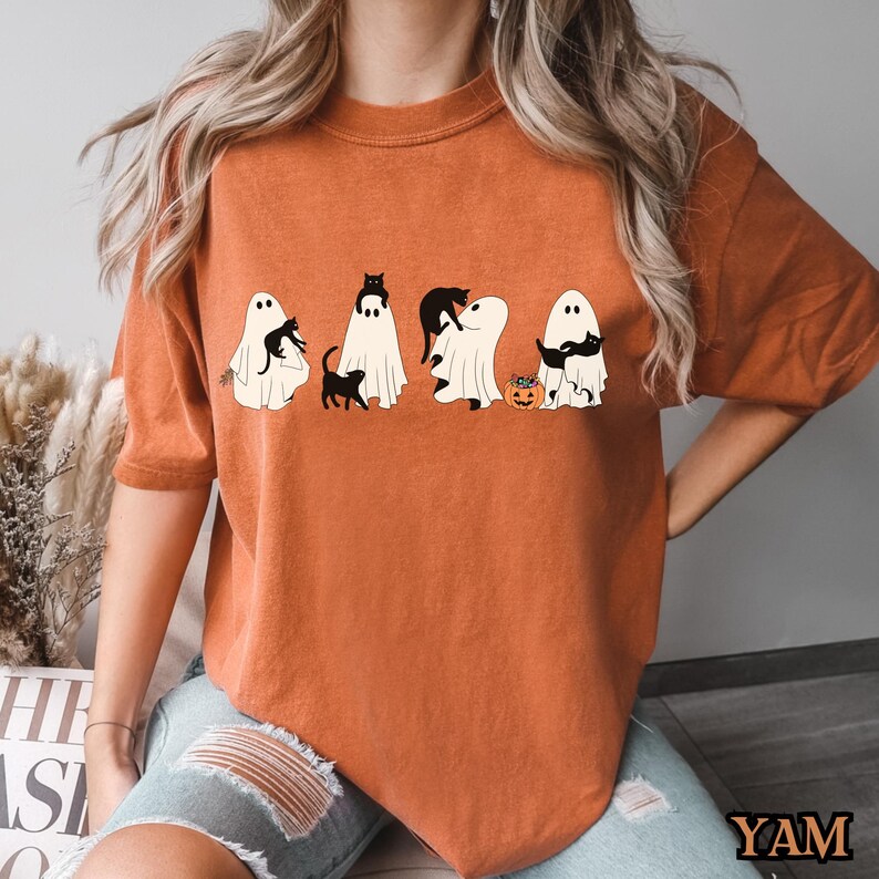 K&ouml;nnte beinhalten: Eine Frau tr&auml;gt ein orangefarbenes T-Shirt mit einem Aufdruck von f&uuml;nf Geistern mit schwarzen Katzen darauf. Die Geister halten K&uuml;rbisse und die Katzen tragen Hexenh&uuml;te. Das Shirt hat einen Used-Look.