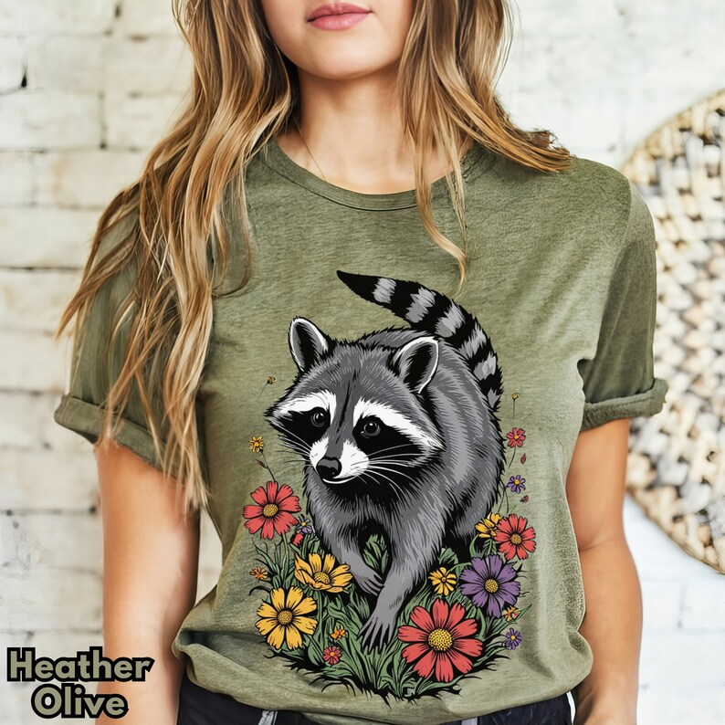 May include: A heather olive green t-shirt with a graphic of a raccoon in a field of flowers. The raccoon is gray and black with a white face and black eyes. The flowers are yellow, red, and purple.
