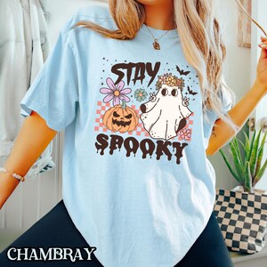 K&ouml;nnte beinhalten: Hellblaues T-Shirt mit einem Grafikdruck eines Geistes, der eine Blumenkrone und einen K&uuml;rbis tr&auml;gt. Der Text "Stay Spooky" ist in Schwarz unter dem Grafikdruck aufgedruckt.