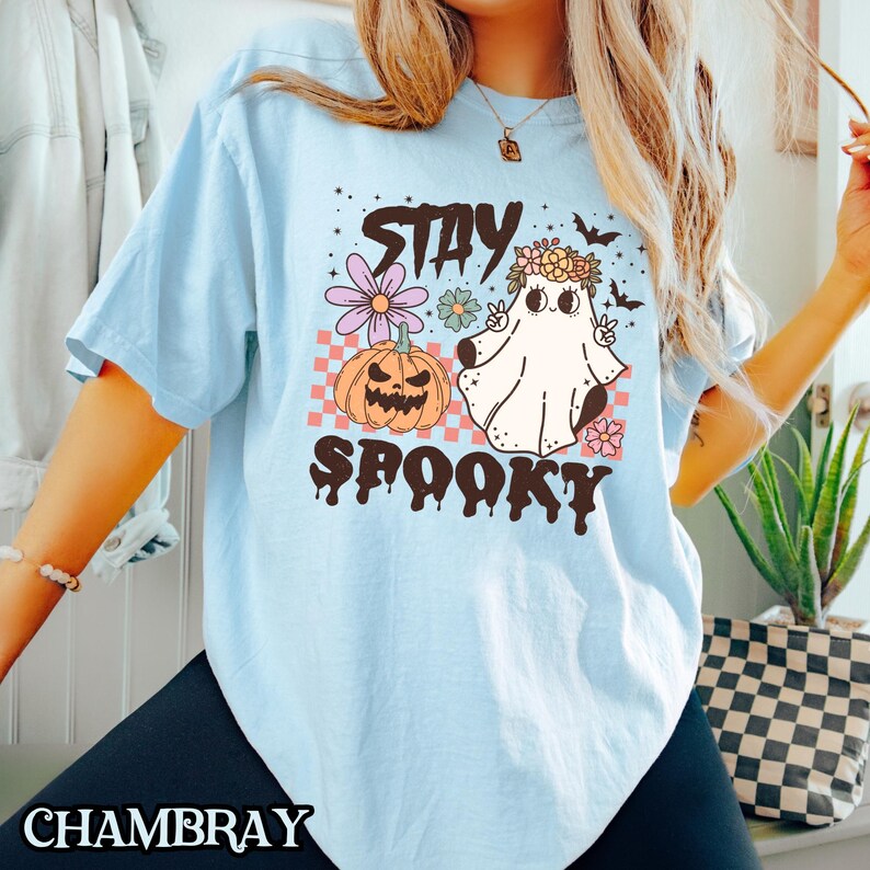 K&ouml;nnte beinhalten: Hellblaues T-Shirt mit einem Grafikdruck eines Geistes, der eine Blumenkrone und einen K&uuml;rbis tr&auml;gt. Der Text "Stay Spooky" ist in Schwarz unter dem Grafikdruck aufgedruckt.