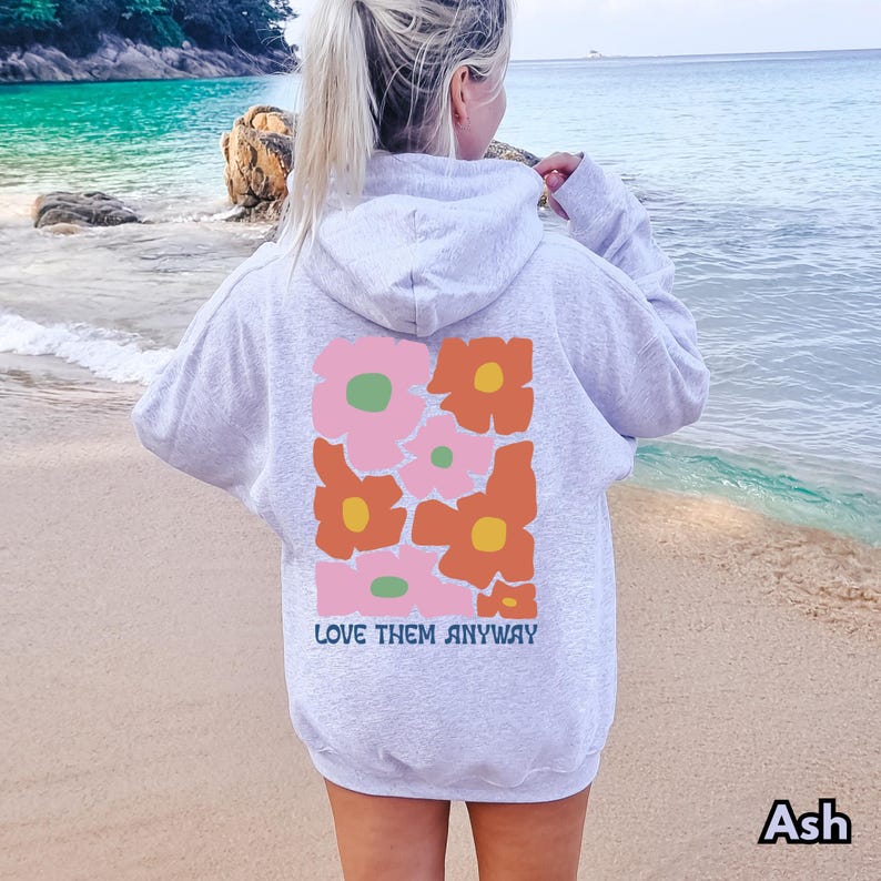 May include: An ash gray hooded sweatshirt featuring a large rectangular graphic on the back. The design displays a pattern of stylized pink and orange flowers with yellow and green centers. The text "LOVE THEM ANYWAY" is printed in blue below.