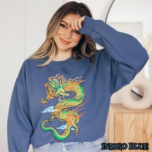 K&ouml;nnte beinhalten: Ein blaues Sweatshirt mit einem gr&uuml;nen und gelben Drachen-Motiv. Der Drache ist von orangefarbenen Flammen und wei&szlig;en Wolken umgeben. Der Text "INDIGO BLUE" ist unten auf dem Sweatshirt aufgedruckt.