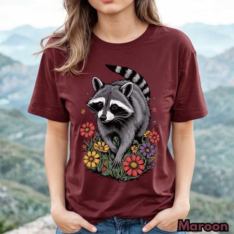 May include: A maroon t-shirt with a graphic of a raccoon walking through a field of colorful flowers.