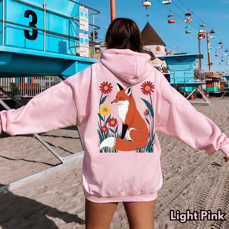 May include: Light pink hoodie with a fox and floral graphic on the back. The fox is orange and white, surrounded by red and yellow flowers. The text "Light Pink" is at the bottom of the image.
