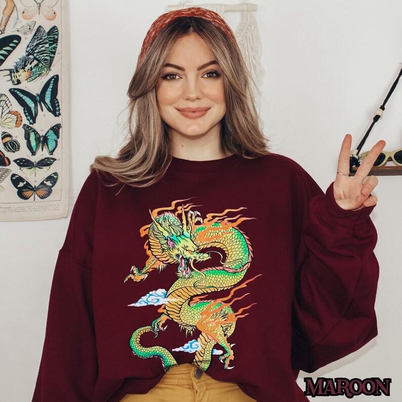 K&ouml;nnte beinhalten: Ein maroonfarbener Pullover mit einem bunten Drachen-Motiv und dem Wort "MAROON" unten rechts.
