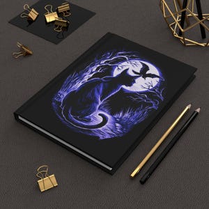May include: A black notebook with a purple illustration of a cat and a bird silhouetted against a full moon. The illustration is surrounded by trees and grass.