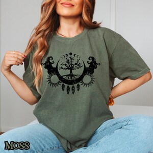 May include: A green t-shirt with a black graphic of a tree with roots, surrounded by moons and suns. The graphic also includes two stylized figures on either side of the tree.