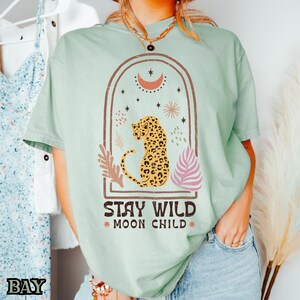 May include: A light green short-sleeve t-shirt featuring a graphic of a leopard inside an arched frame with celestial elements. Below the leopard, the text reads STAY WILD MOON CHILD in dark brown letters, accented by small star and sun symbols.