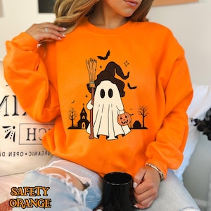 以下が含まれることがあります： オレンジ色のスウェットシャツに、魔女の帽子をかぶってほうきとカボチャを持った幽霊のグラフィックがプリントされています。幽霊は教会と木の前にいます。