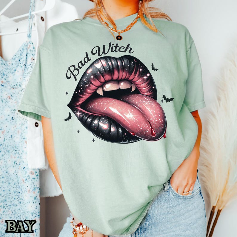 Peut inclure: Un t-shirt vert clair avec le texte "Bad Witch" et un graphique d'une bouche ouverte noire et rose avec une langue qui sort.