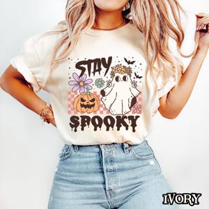 K&ouml;nnte beinhalten: Ein wei&szlig;es T-Shirt mit einem Aufdruck eines Geisters, der eine Blumenkrone tr&auml;gt und ein Peace-Zeichen macht. Der Geist befindet sich neben einem K&uuml;rbis und der Text "Stay Spooky" befindet sich unter dem Aufdruck.
