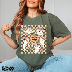May include: A green t-shirt with a checkered pattern featuring a pumpkin and five ghosts wearing hats. The ghosts are white with pink and purple accents. The pumpkin is orange with a black face.