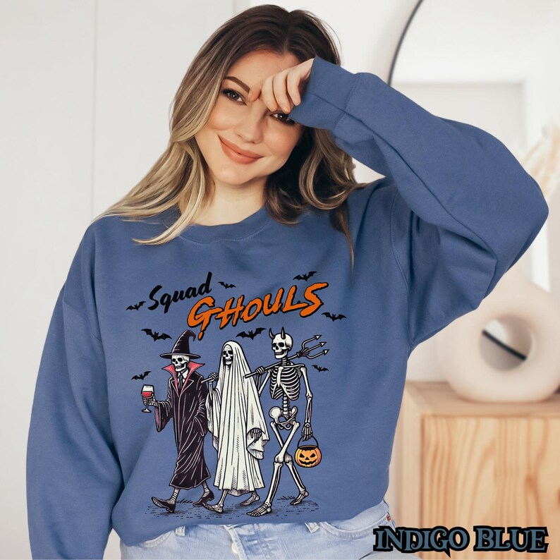 Pu&ograve; includere: Una felpa blu con una grafica di tre scheletri vestiti da strega, fantasma e diavolo. Il testo "Squad Ghouls" &egrave; sopra gli scheletri.