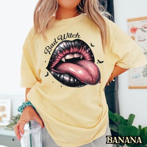 Peut inclure: Un t-shirt jaune avec le texte "Bad Witch" et un graphique d'une bouche noire et rose avec des crocs et une langue.