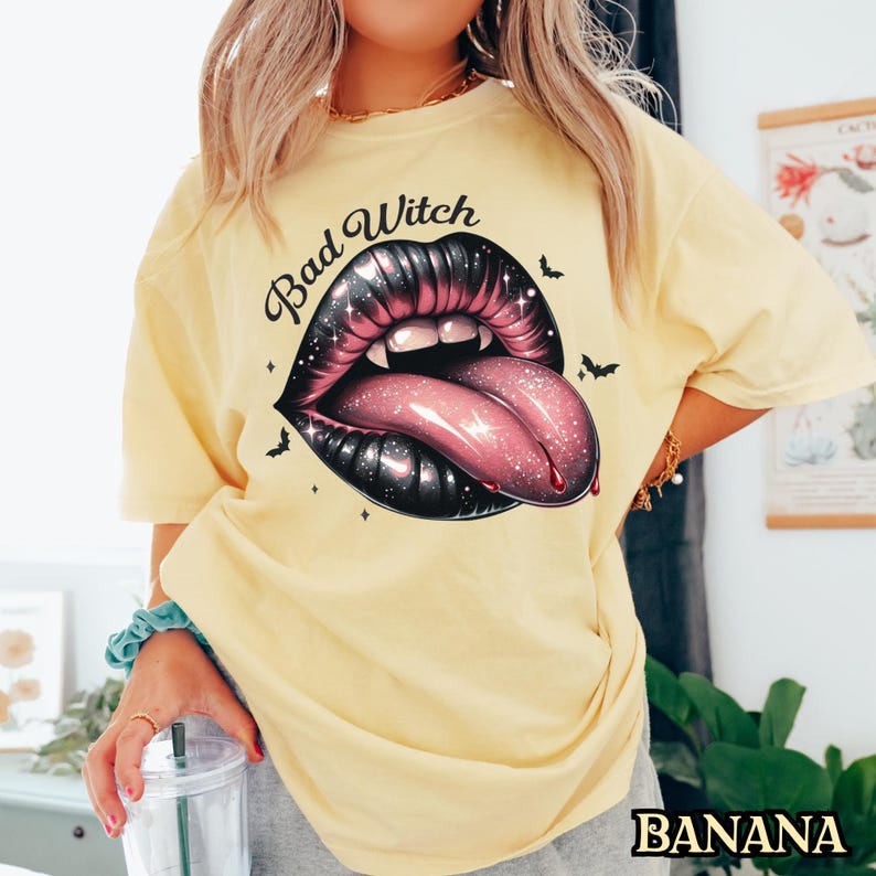 Peut inclure: Un t-shirt jaune avec le texte "Bad Witch" et un graphique d'une bouche noire et rose avec des crocs et une langue.