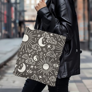May include: A tote bag with a black and white celestial pattern. The pattern includes moons, stars, clouds, suns, and a yin yang symbol. The bag is made of a canvas material and has a black handle.