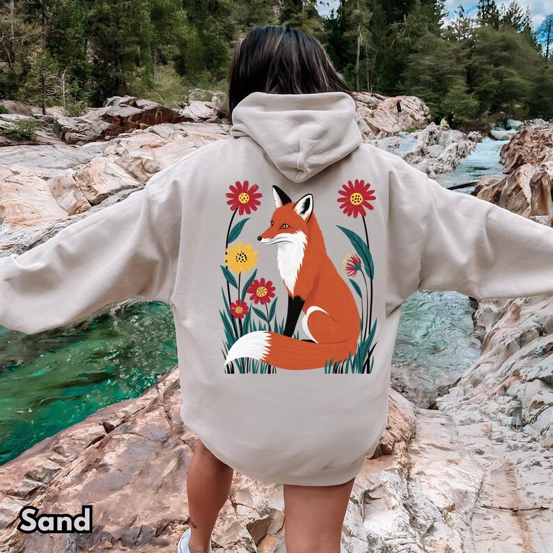 May include: Sand-colored hoodie featuring a vibrant illustration of a fox amidst colorful flowers. The fox is depicted in shades of orange, white, and black, surrounded by red and yellow floral accents. The hoodie has a drawstring hood and a kangaroo pocket.